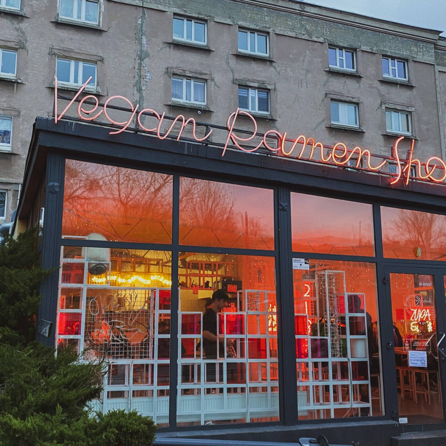 Der Vegan Ramen Shop sticht mit seinem offenen Ambiente und der bunten, leuchtenden Inneneinrichtung vor grauer Häuserkulisse deutlich heraus.