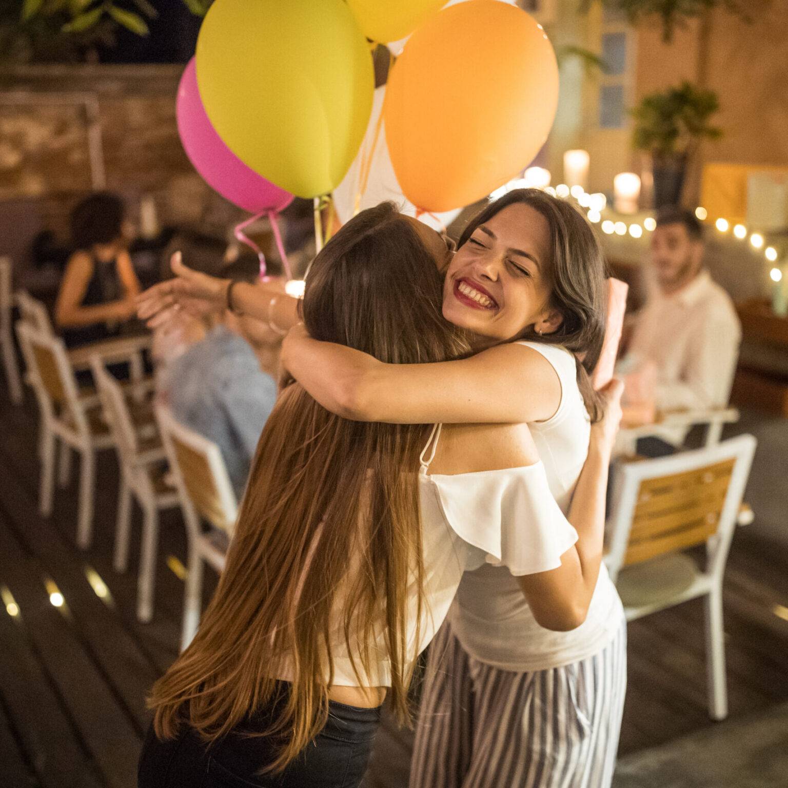 Zwei Frauen umarmen sich. Im Hintergrund sitzten die Gäste der Geburtstagsfeier an einem Tisch.