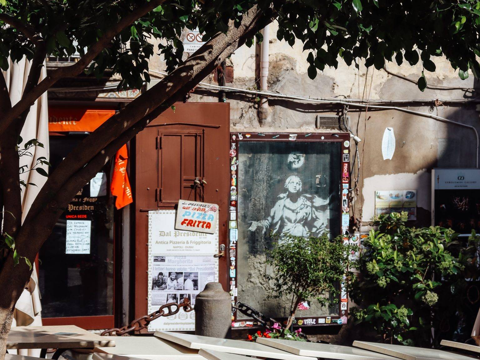 Auf dem Bild von Banksy sehen wir Madonna mit einer Pistole, davor stehen Kaffeetische und ein Baum.