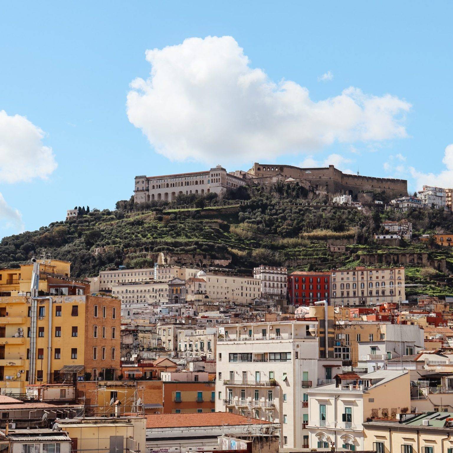 Ein imposanter Blick über viele Häuserdächer Neapels, dahinter eine Erhebung mit dem mediterranen Schloss Castel Sant'Elmo.