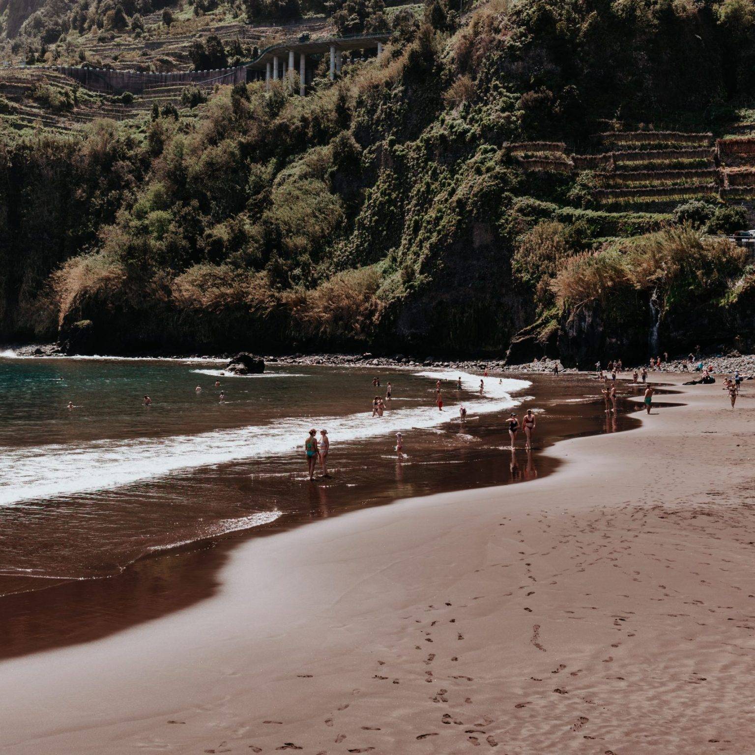 Ein flacher Strand in einer grünen Bucht: Links türkises Wasser, rechts dunkler Sand.