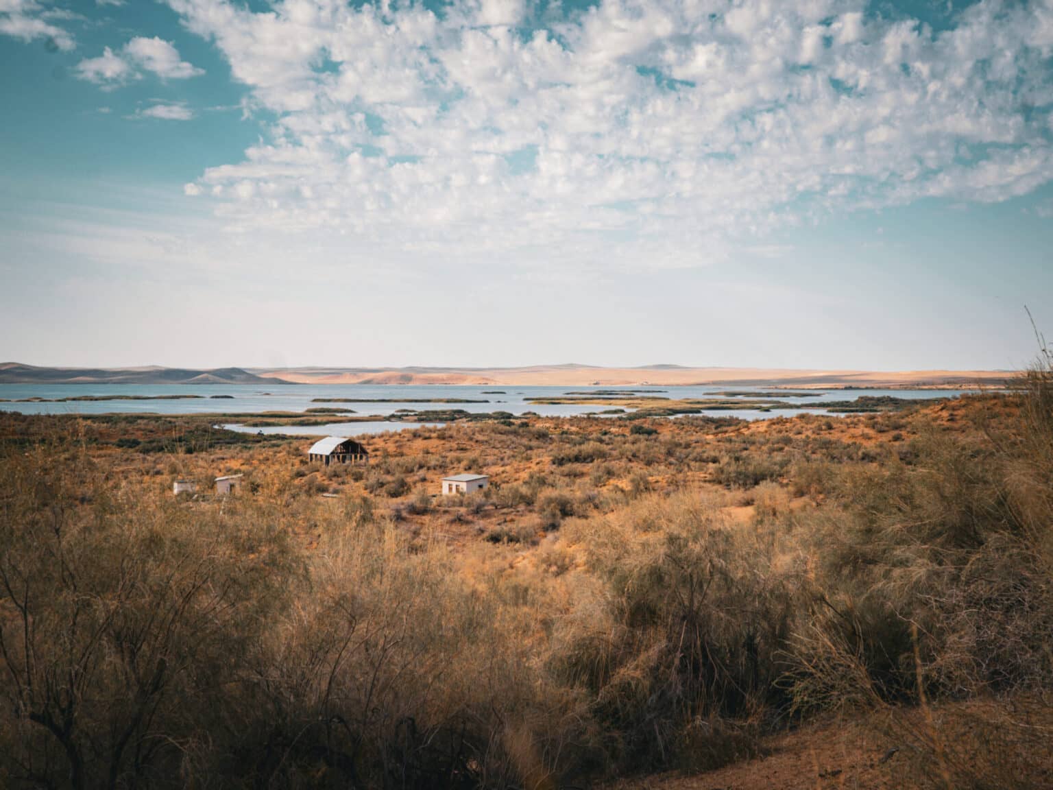 Blick auf den Aydarkul-See in Usbekistan, umgeben von trockener Wüstenlandschaft und vereinzelten Hütten