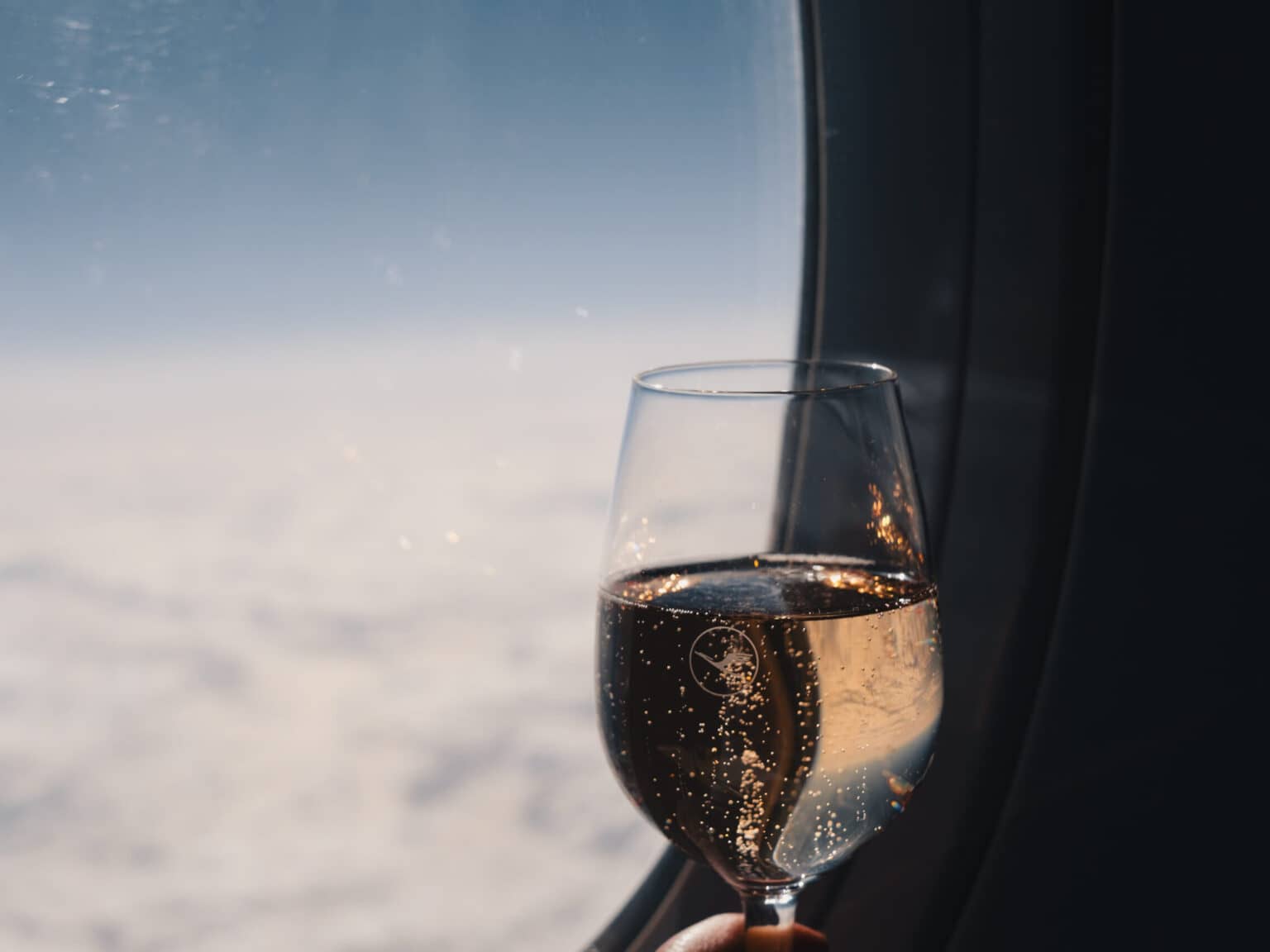 Ein Glas Champagner vor einem Flugzeugfenster. Im Hintergrund ist eine weiße Wolkendecke und darüber blauer Himmel.