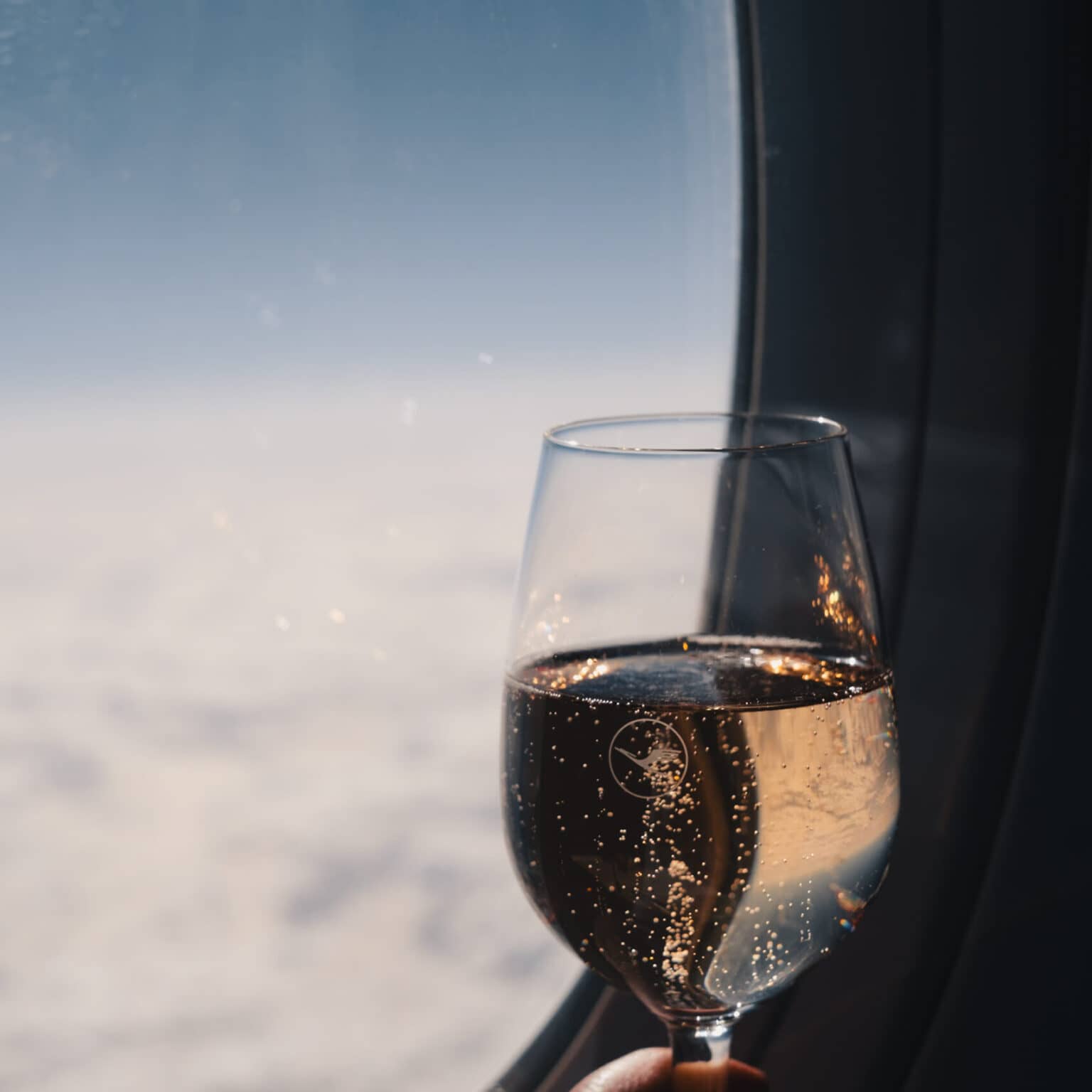 Ein Glas Champagner vor einem Flugzeugfenster. Im Hintergrund ist eine weiße Wolkendecke und darüber blauer Himmel.