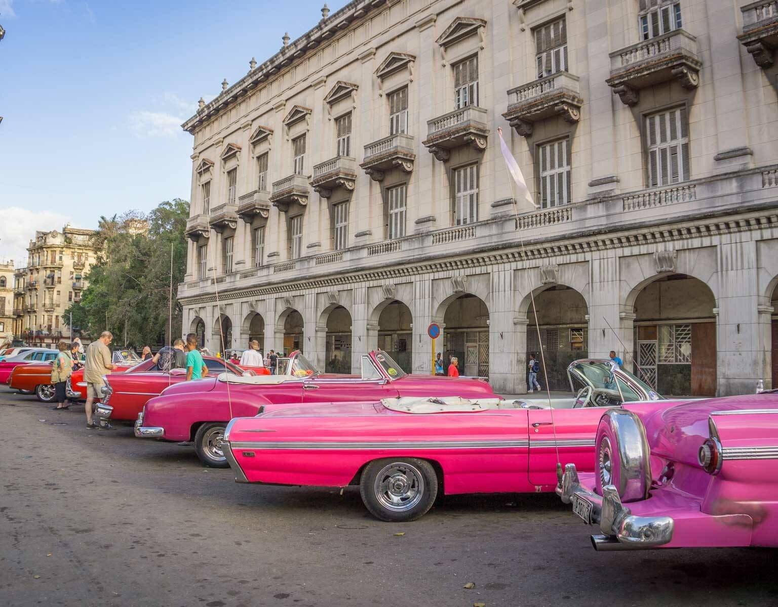 Mehrere Oldtimer in unterschiedlichen Rottönen stehen vor einem Häuserzug in Havanna, Kuba.