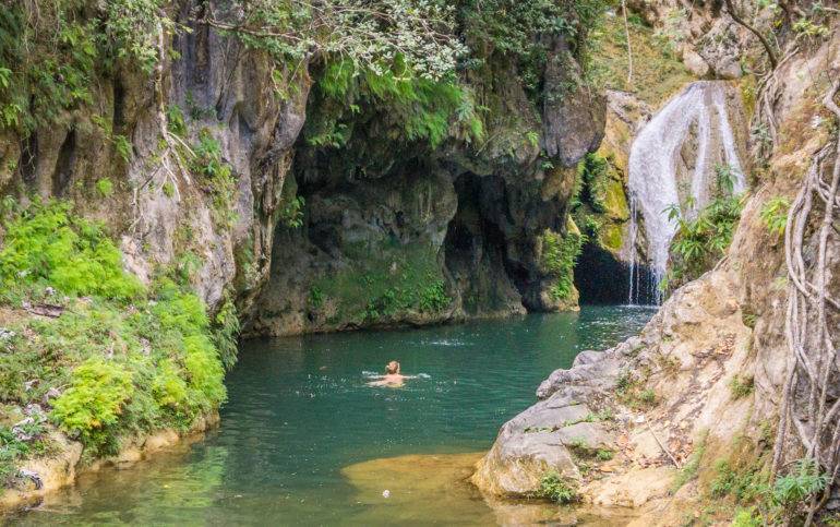 In Trinidad, Kuba, befindet sich im Parque el Cubano zwischen bewachsenen Felsvorsprüngen ein Wasserfall zu dem eine Frau schwimmt.