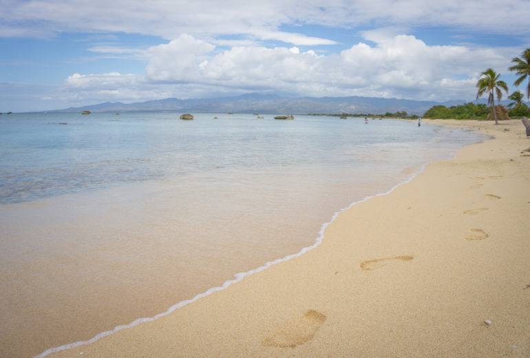 An einem menschenleeren Sandstrand von Kuba sind Fußabdrücke im Sand zu sehen während das türkisfarbene Wasser sanft ans Ufer rollt.