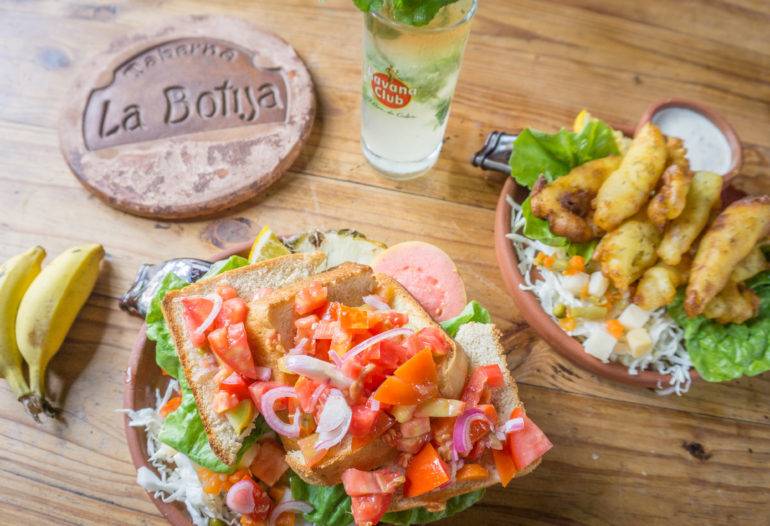 Auf dem Holztisch der Taberna La Bojita in Trinidad, Kuba sind zwei Teller mit Salat und Toast, sowie einem Mojito und Babybananen angerichtet.