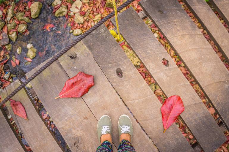 Zwei Füße stehen auf einer kleinen hölzernen Hängebrücke über rotgelbem Blättermeer in Kubas Parque el Cubano.