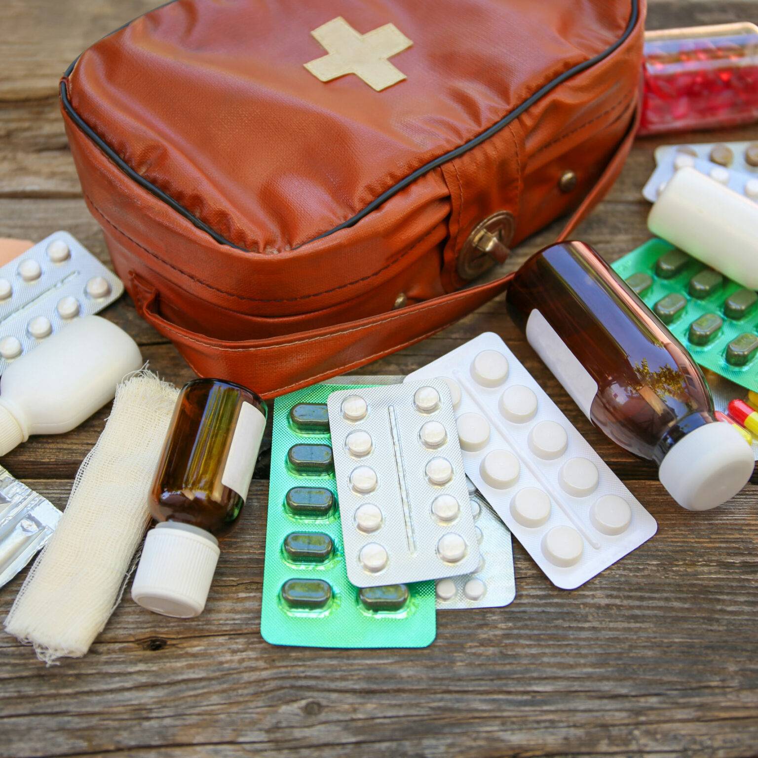 Eine Reiseapotheke in Form einer Erste-Hilfe-Tasche liegt auf Holz, davor liegen Tabletten, Zäpfchen, Medikamentenflaschen und Verbandszeug.