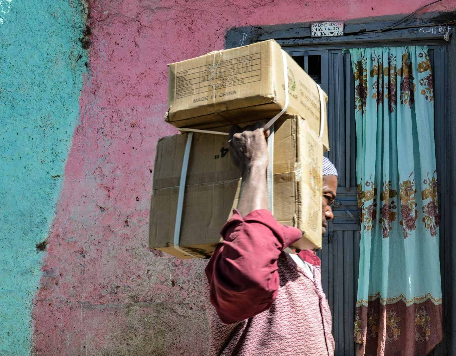 In Harar, Äthiopien, trägt ein dunkelhäutiger Mann vor einer blau-lila Häuserwand zwei Pakete auf der Schulter durch die Straße.