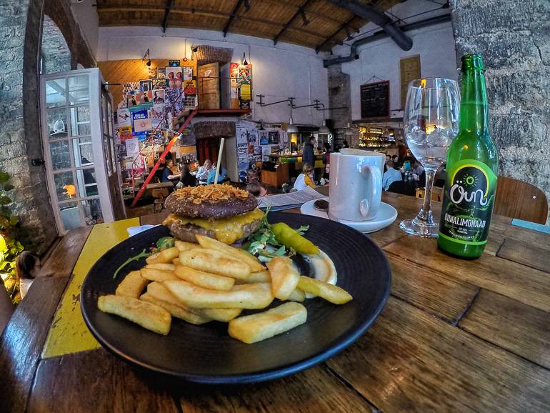 In Tallinns Restaurant F-hoone wird an einem Holztisch Burger mit Pommes serviert, im Hintergrund ist eine Wand mit bunten Postern zu sehen.