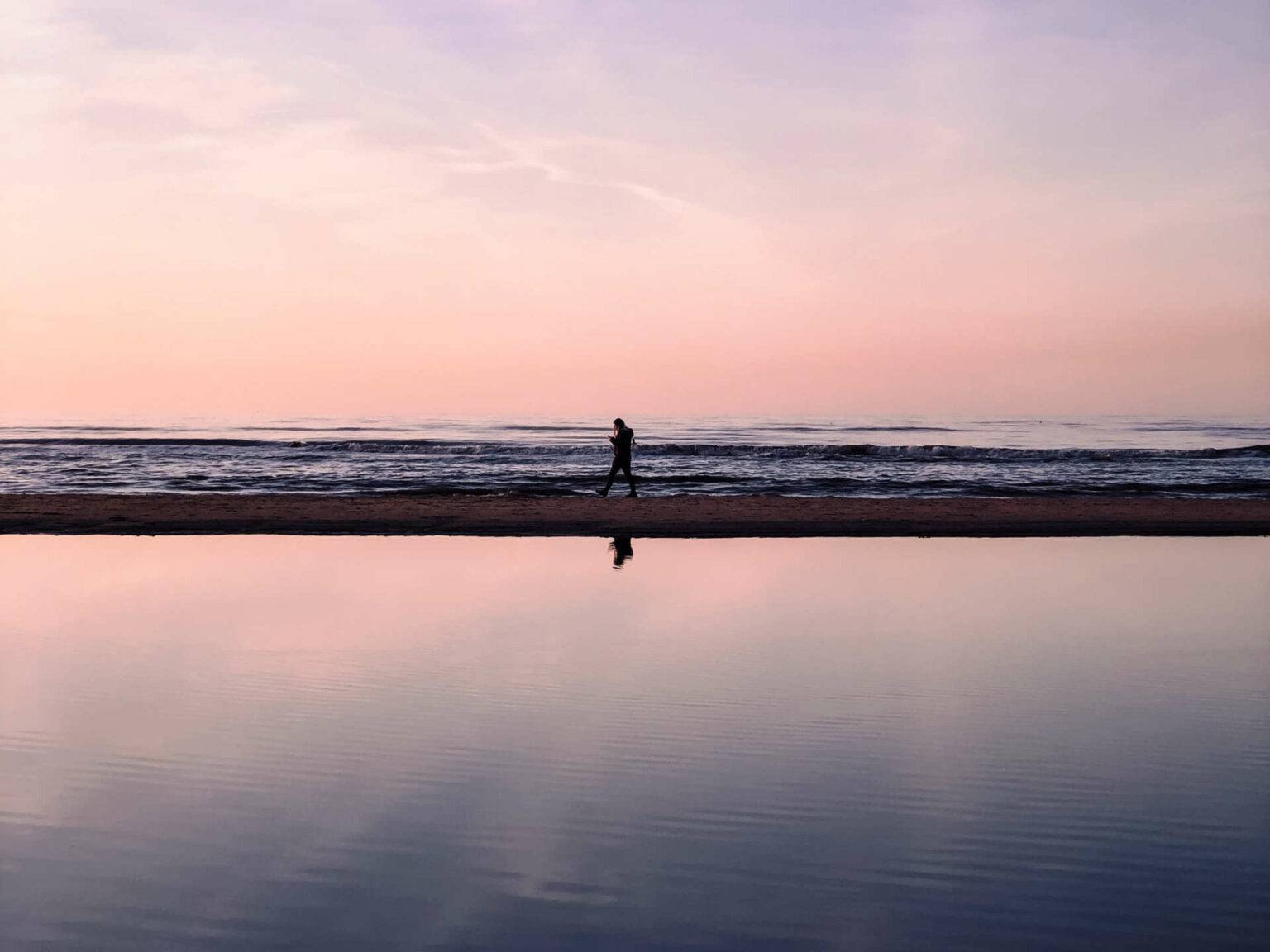 Lila und pink gefärbter Himmel, darunter das Meer und eine kaum erkennbare Person, die den Strand entlang spaziert