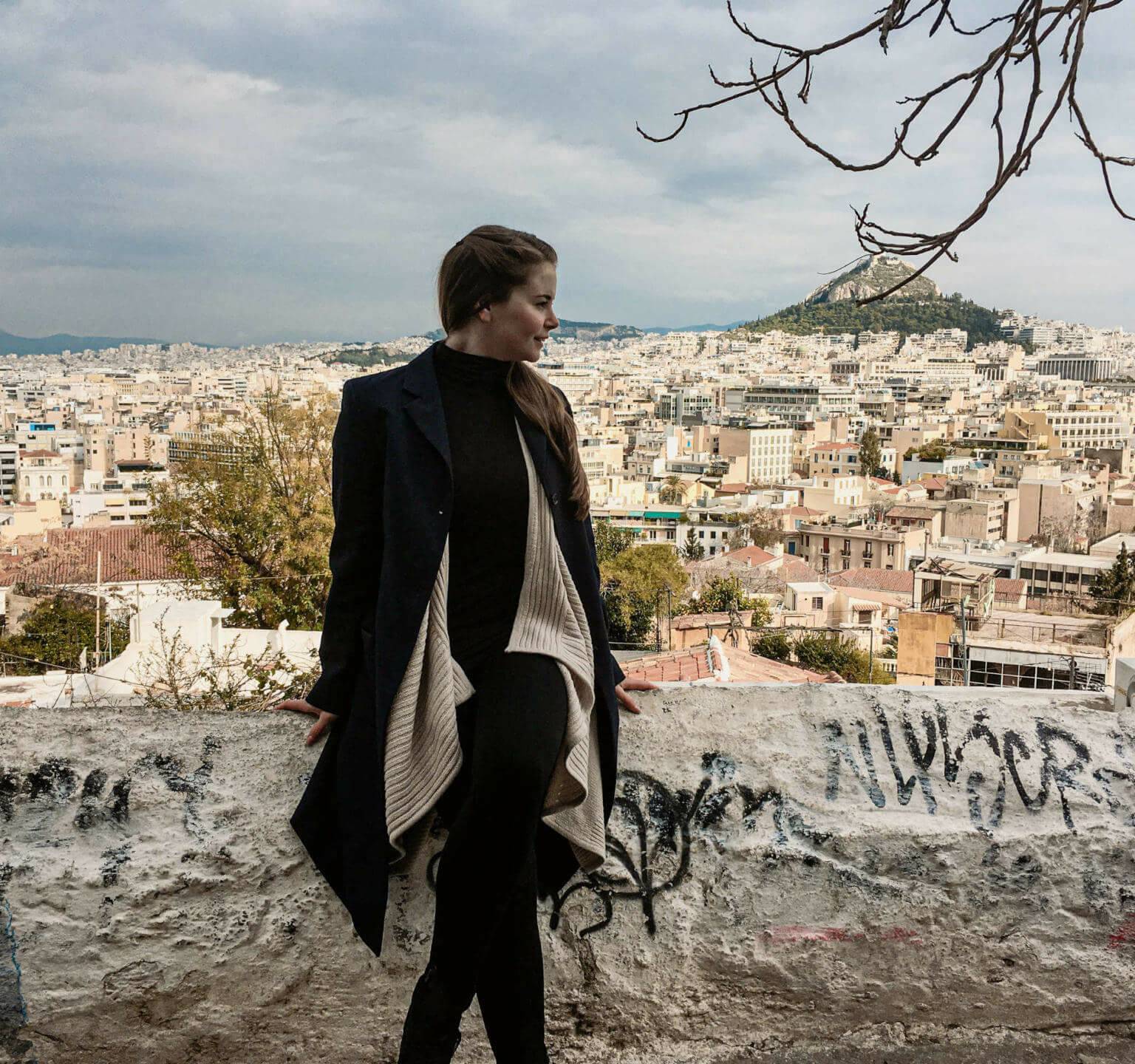 Blick auf die sich weit erstreckende Hauptstadt Athen und die Akropolis von Anafiotika aus, Reisebloggerin Franziska posiert vor der Dächerflut auf einer Mauer sitzend.