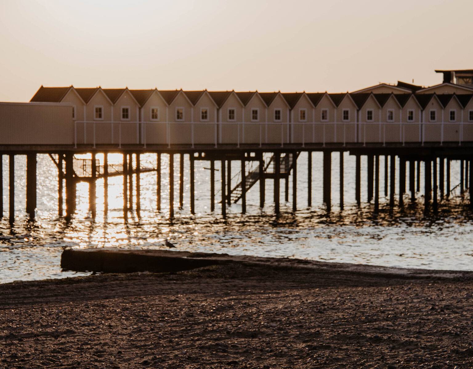 Eine lange Reihe von Häusern auf Stelzen, in denen sich Kaltbadehäuser befinden, liegen ein hohes Stück über dem seichten Wasser in der Abenddämmuerng, der Sand vom Strand färbt sich dunkel im Licht, eine Treppe aus dem Wasser führt die weißen Häuser hinauf.