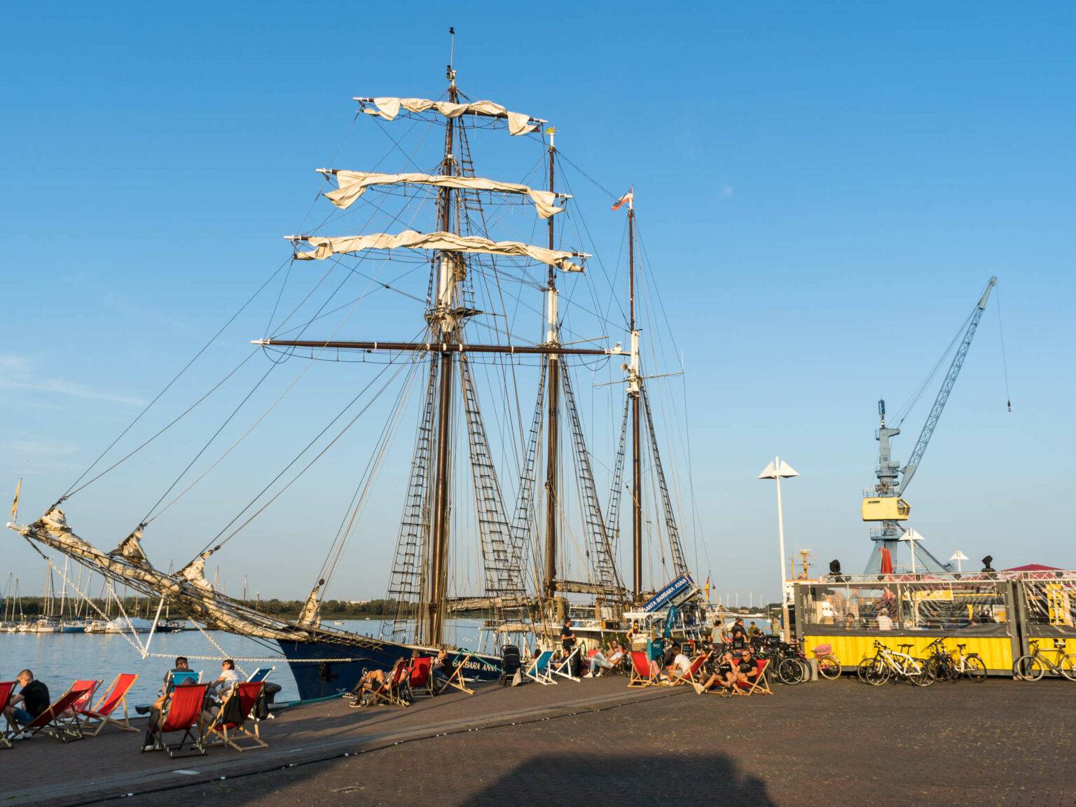 Ein umfunktioniertes Schiff liegt in Rostock an einem Dock an, davor sitzen Menschen in Strandstühlen, daneben befindet sich ein kleiner Unterstand mit anlehnenden Fahrrädern, der Himmel ist strahlend blau, man blickt auf einen Kran.
