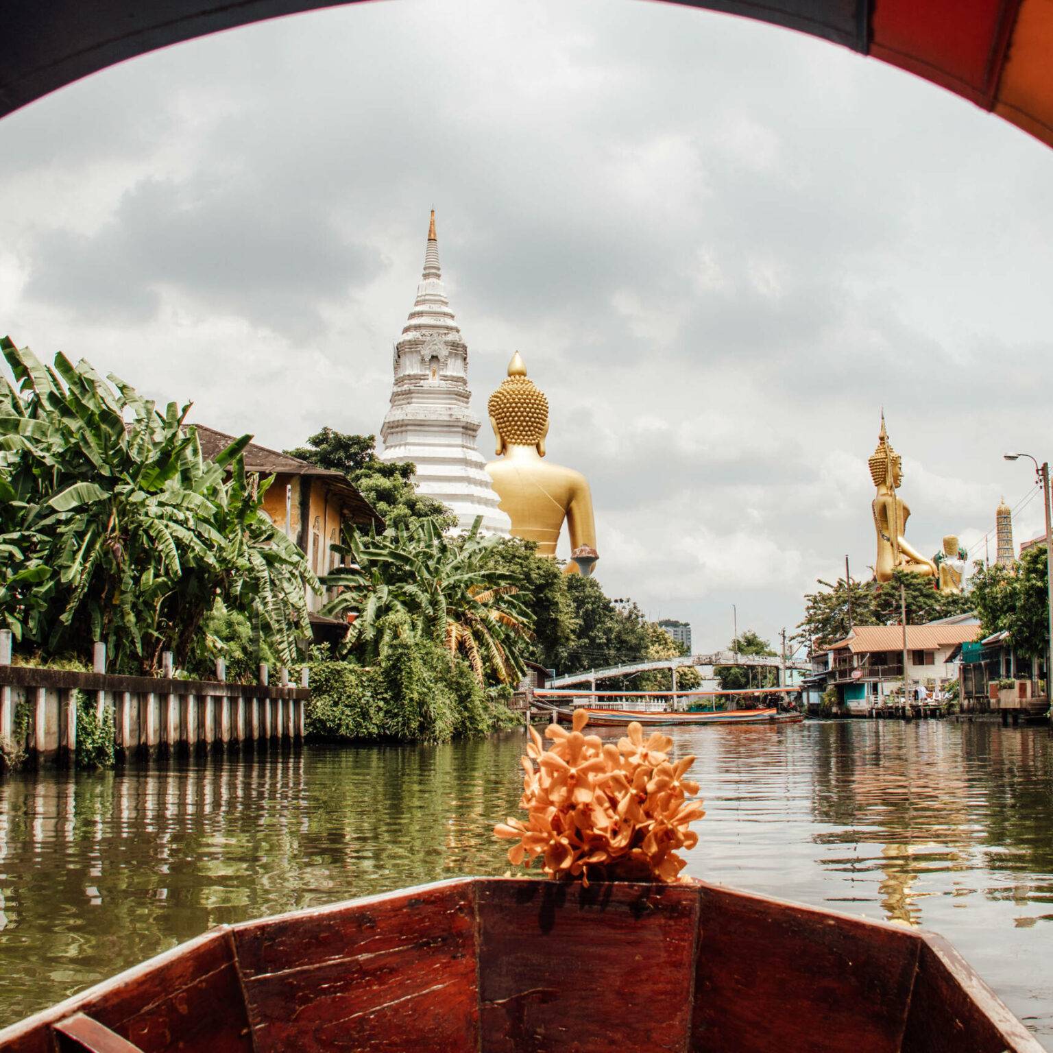 Blick von einem Boot auf einem breiten Fluss in Bangkok mit goldenen Skulpturen im Hintergrund.