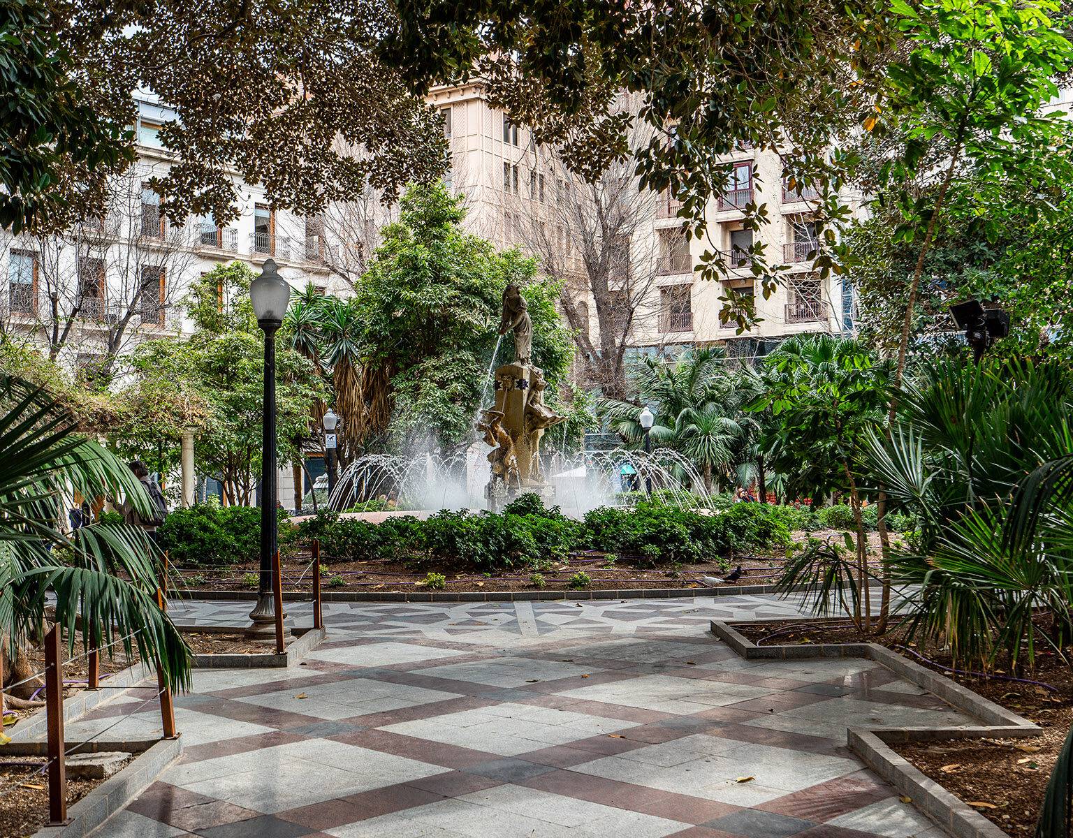 Ein Brunnen inmitten der Altstadt von Alicante zwischen vielen Bäumen und Palmen.