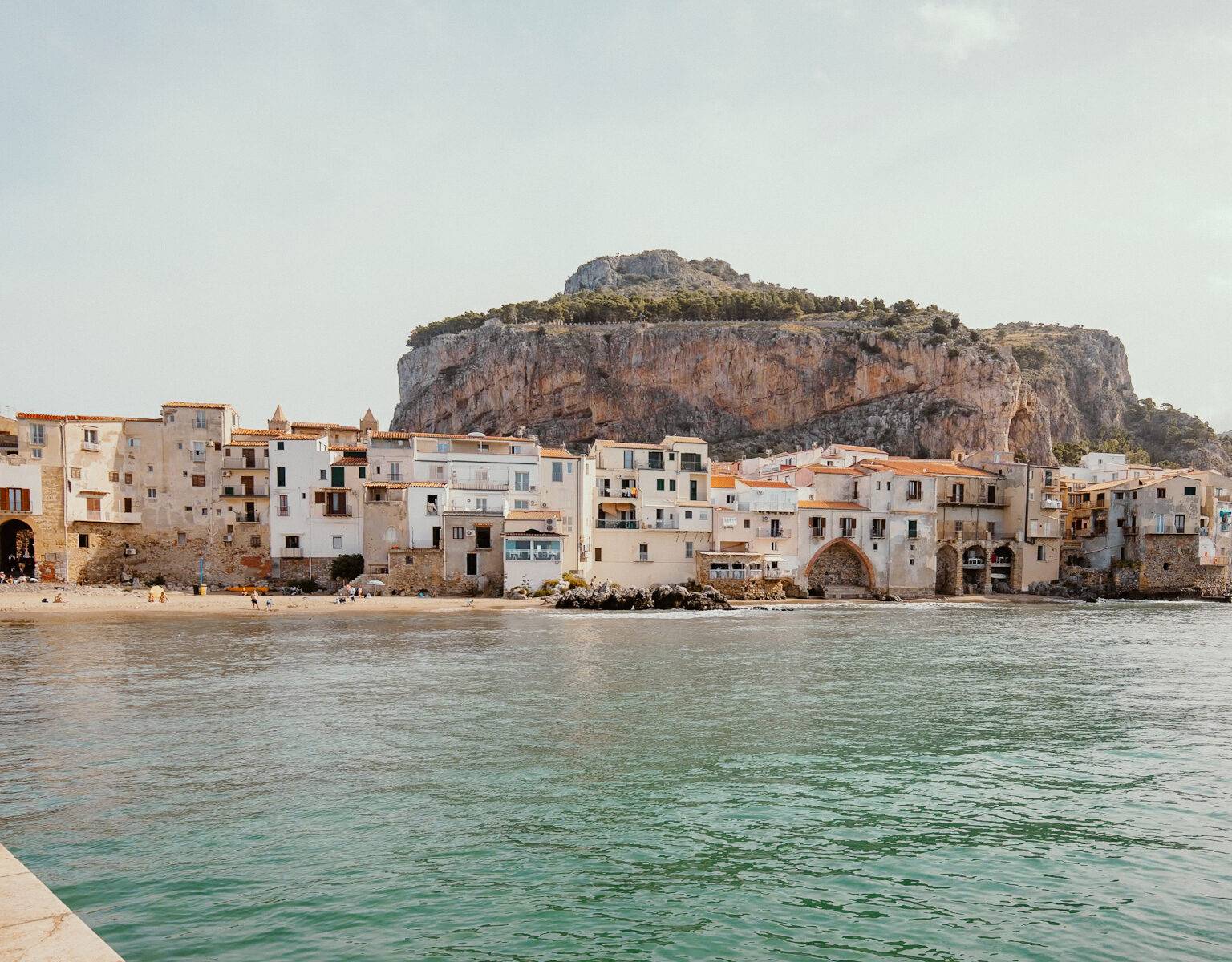 Ein Stadtstrand in Cefalu, im Hintergrund erstreckt sich ein Felsen über die Stadt.