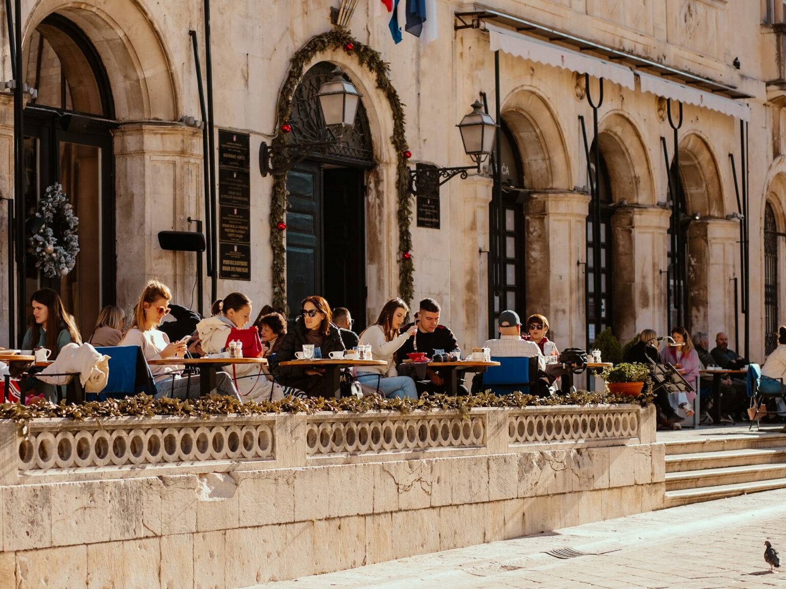 Menschen sitzen auf der Terrasse des Gradska Kavana Arsenal Restaurants in der Sonne.