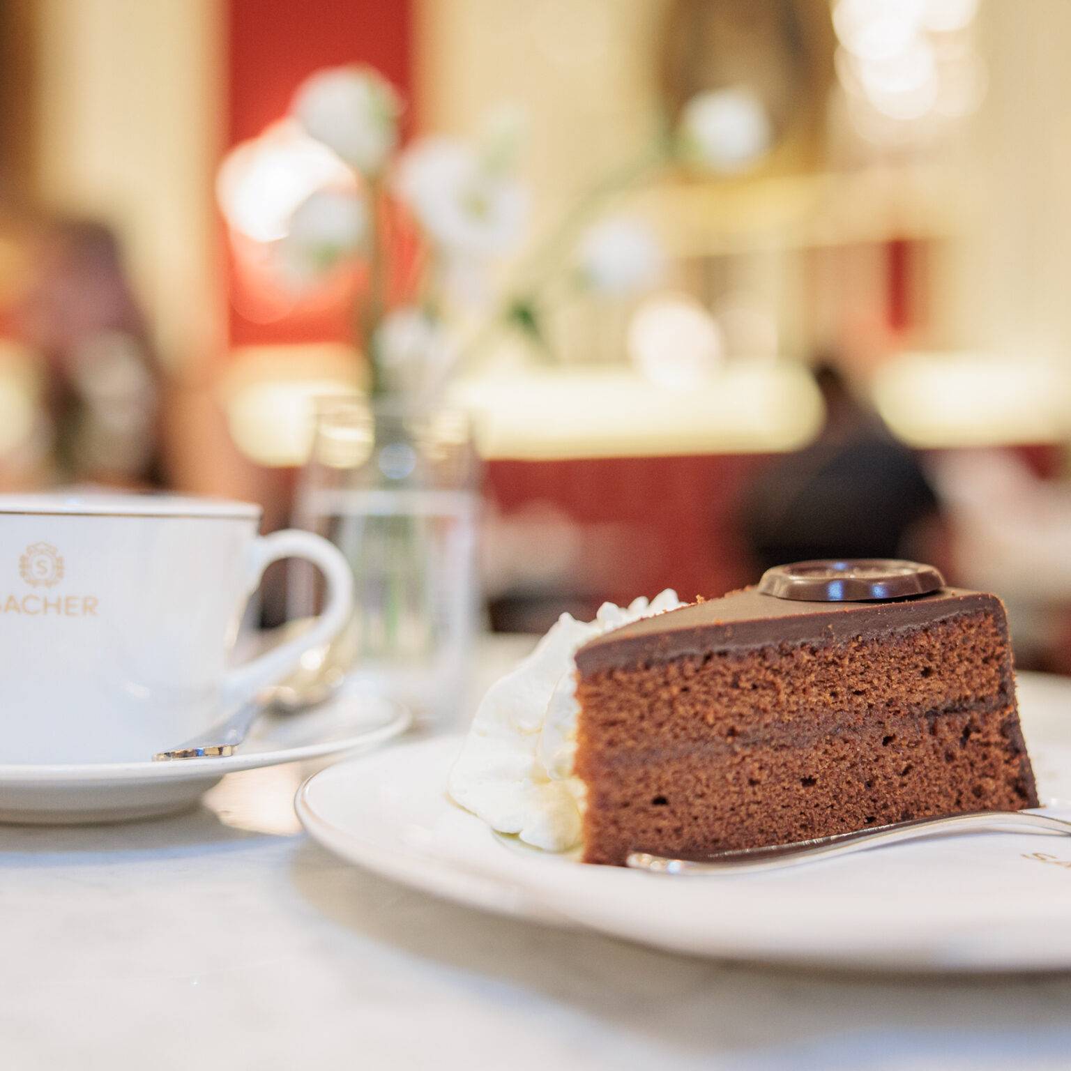 Tasse Kaffee im Kaffeehaus Sacher neben einem Stück Sachertorte auf einem Teller, mit einer Vase Blumen im Hintergrund.