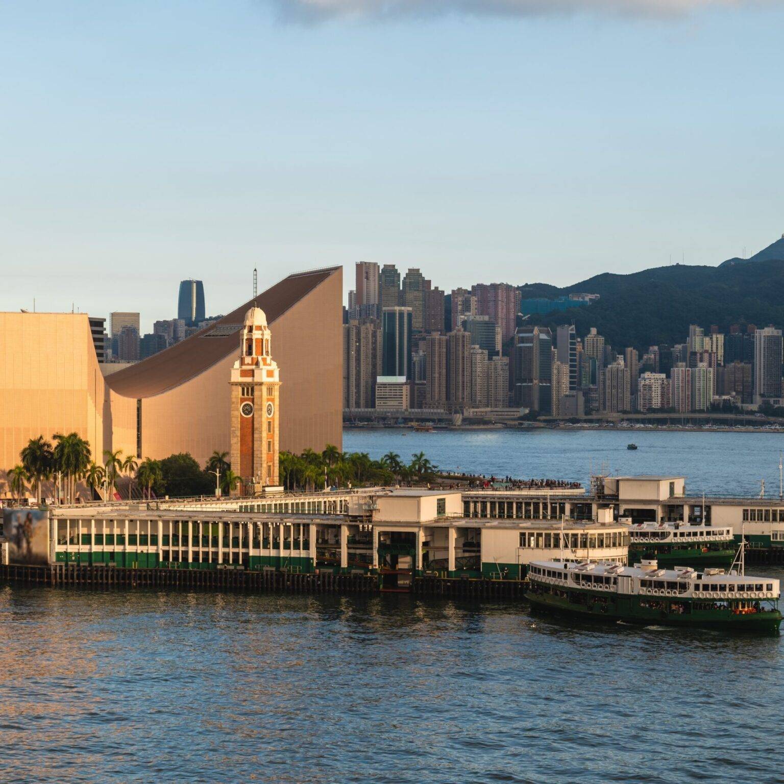 Perspektive vom Wasser auf das Ufer von Tsim Sha Tsui in Hongkong bei Sonnenuntergang mit Fähren und einem Kirchturm.